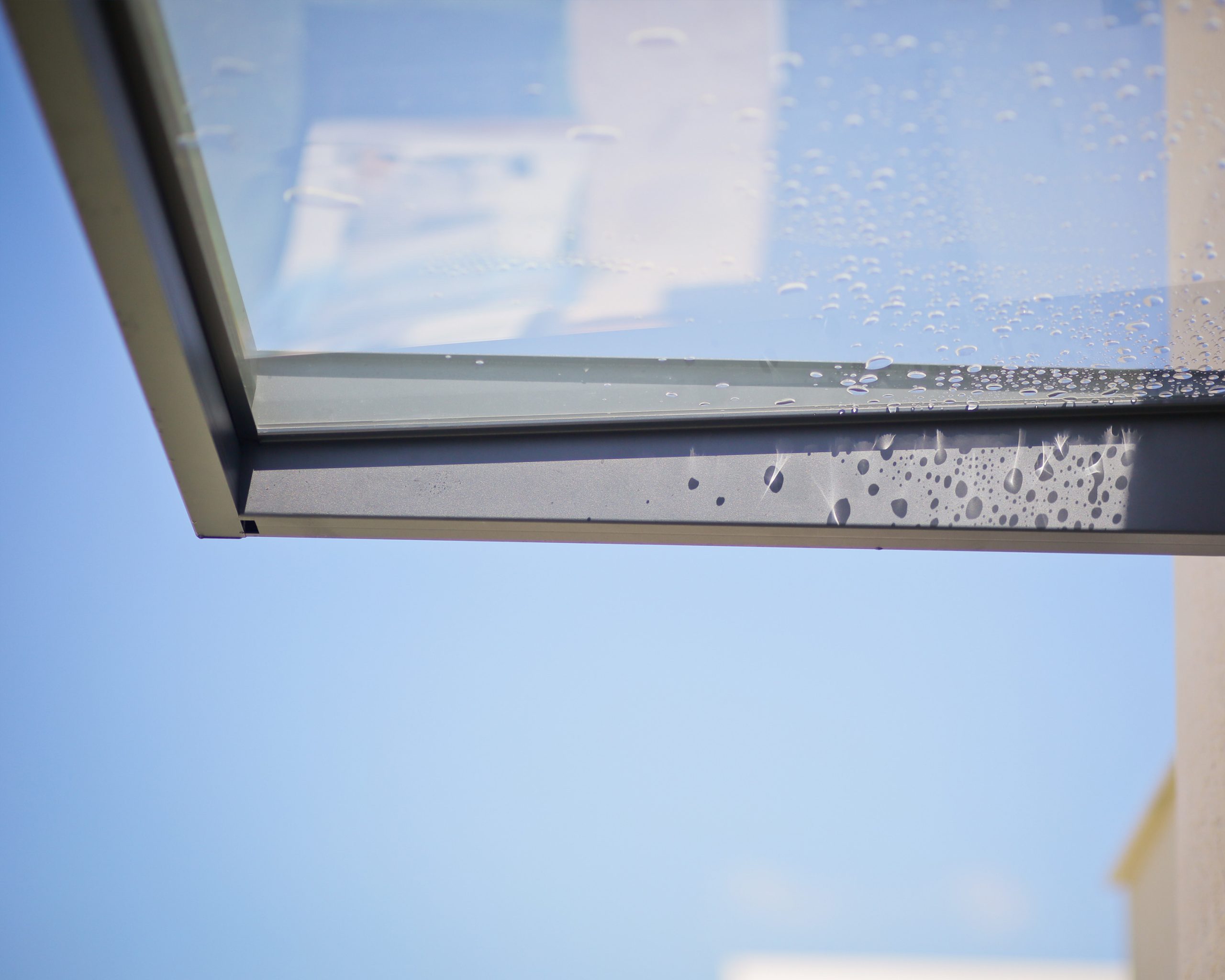 Sophia 2150 door canopy featuring a gray aluminum frame and clear panels, showcasing built-in gutter with water droplets on the surface against a blue sky.