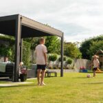 Family enjoying outdoor space under a modern NOVA Titan Aluminium Pergola, featuring a sleek design, children playing on lawn, and comfortable seating area.