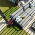 Stylish outdoor seating area featuring grey cushions and decorative pillows, surrounded by greenery and floral accents, under the shade of a modern pergola.